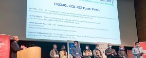 Many researchers and a presenter stand under a projection with the names of the winning posters.